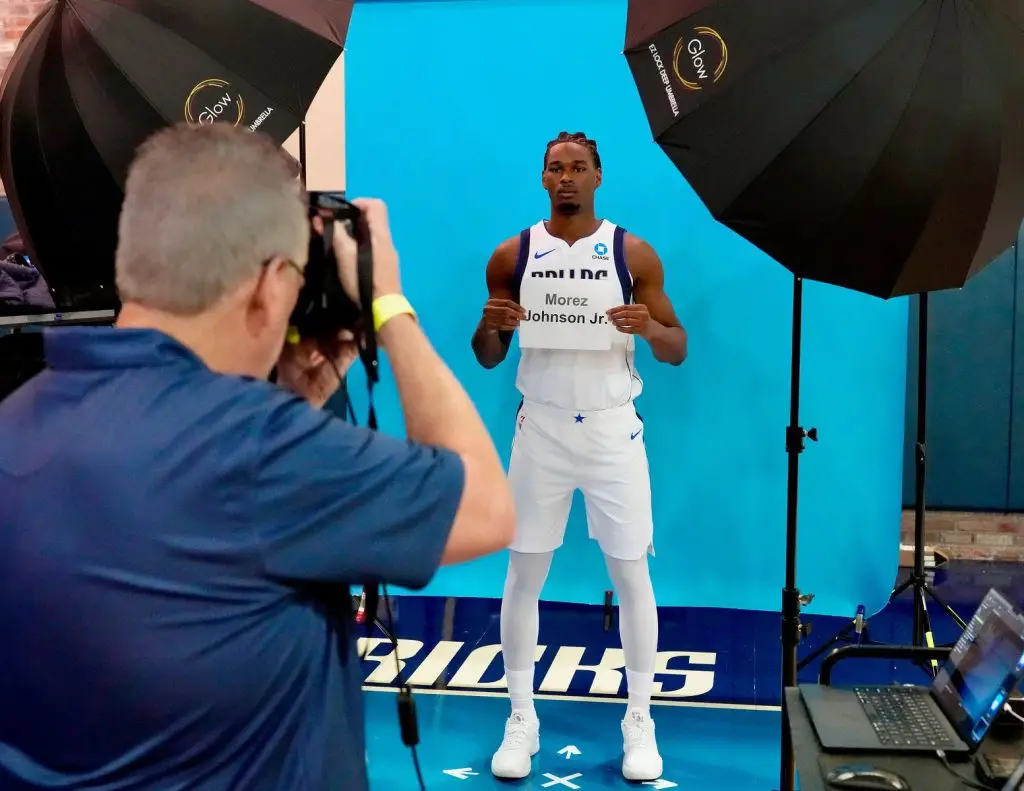 Photographer photographing a player in a white Dallas jersey who holds a name card in front of a blue backdrop between two umbrella lights
