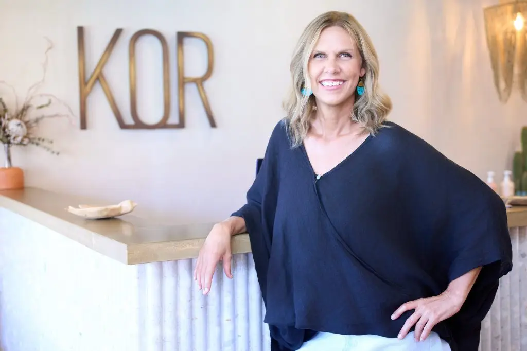 Hair stylist in a black draped top leaning against the reception desk below the wooden KOR wall logo