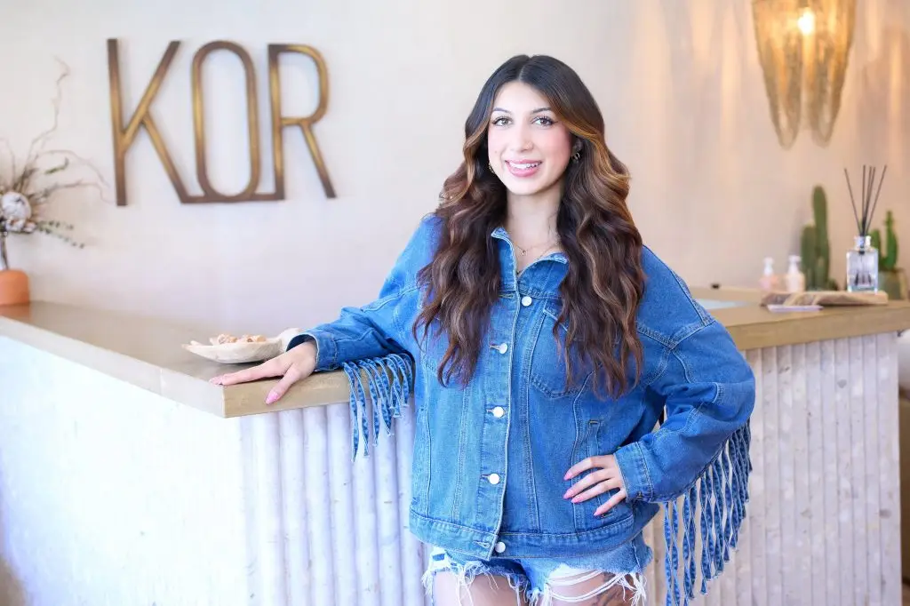Hair stylist in a fringed denim jacket resting one hand on the salon reception counter