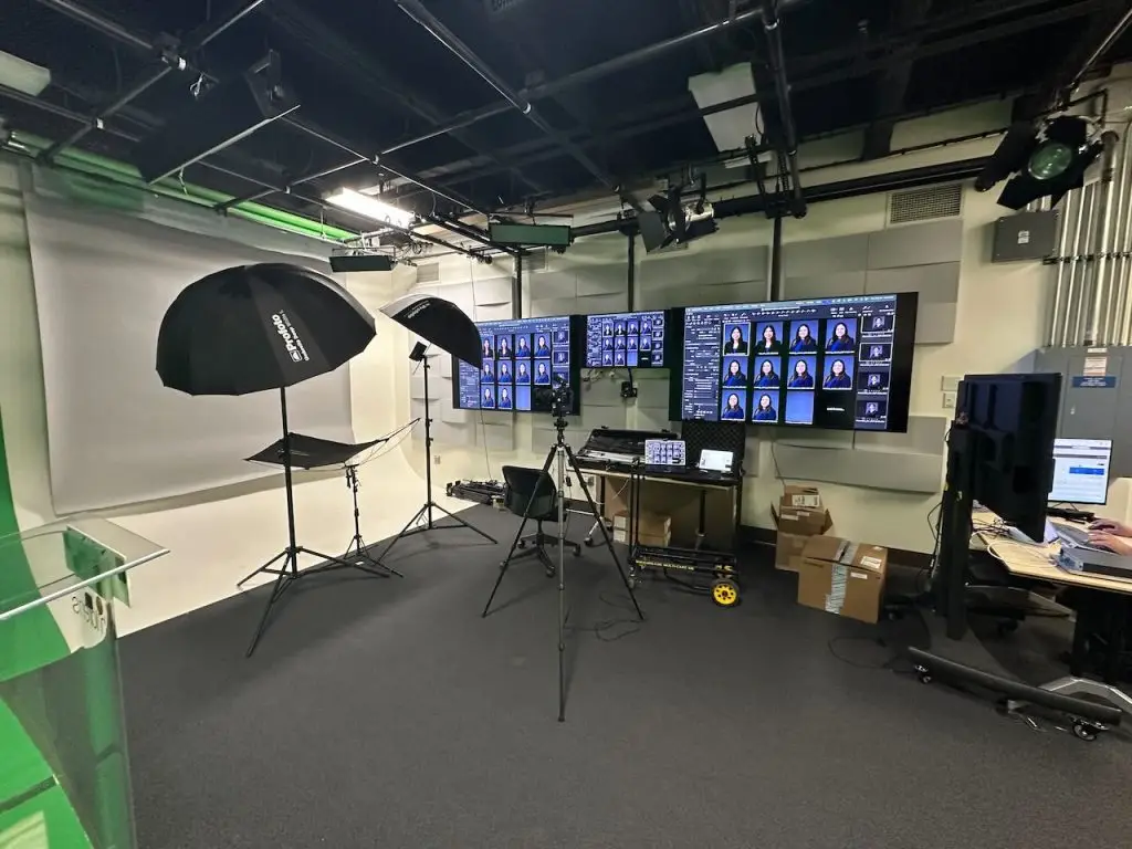 Headshot lighting and a camera on tripods in a studio room, with three large monitors displaying grids of headshots and a computer workstation to the right