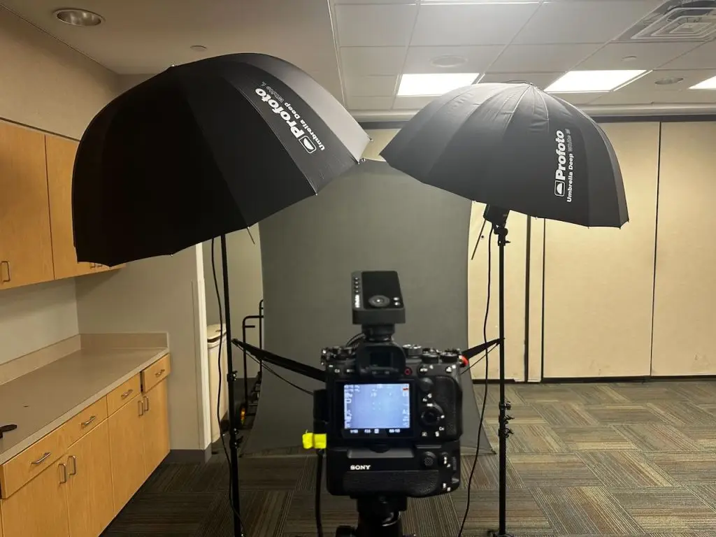 Camera on a tripod facing a gray backdrop lit by two large umbrella lights in a hospital meeting room