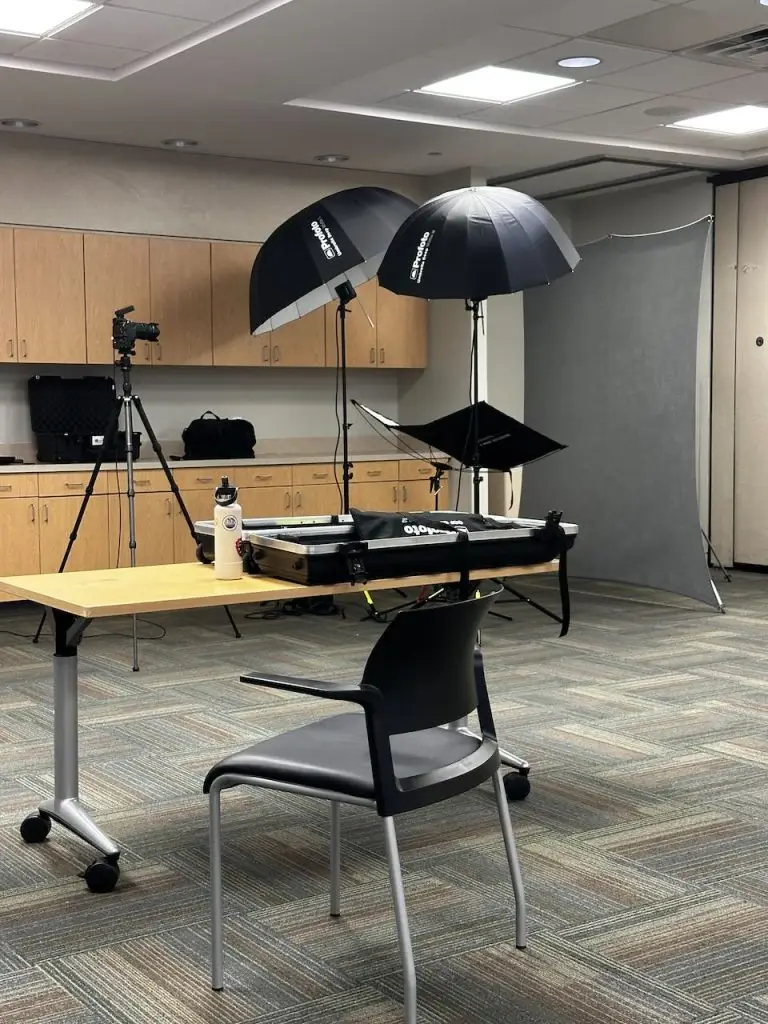 Portable headshot studio with umbrella lights, a gray backdrop and a tripod-mounted camera set up in a meeting room, with a chair and table in the foreground